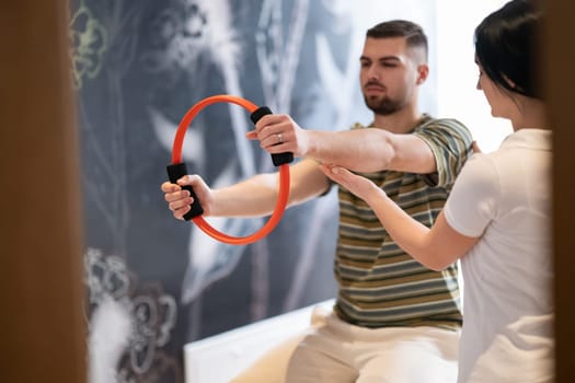 A male patient performing arm exercises using a resistance ring under supervision during a rehabilitation session.