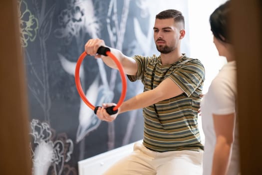 A male patient performing arm exercises using a resistance ring under supervision during a rehabilitation session.