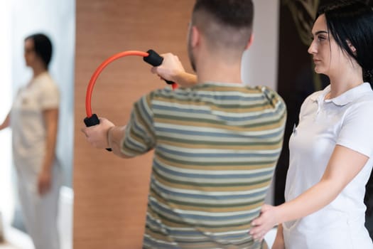 A male patient performing arm exercises using a resistance ring under supervision during a rehabilitation session.