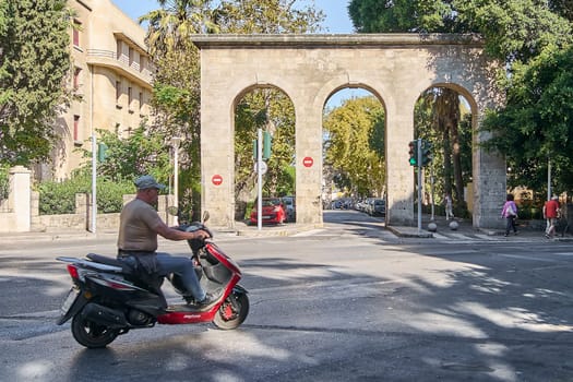 Rodas.Greece - june 04, 2025: A local on a scooter passes through a historic archway in Rhodes town, depicting urban daily life under a clear sky.