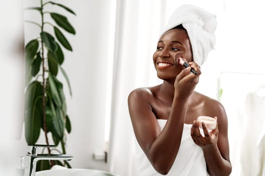 A woman smiles while applying a skincare product to her face in a well-lit bathroom. She wears a towel on her hair and holds a jar in one hand. A plant decorates the space.