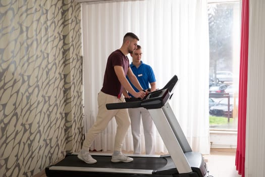 Man undergoing physical therapy with trainer assistance on a treadmill indoors.