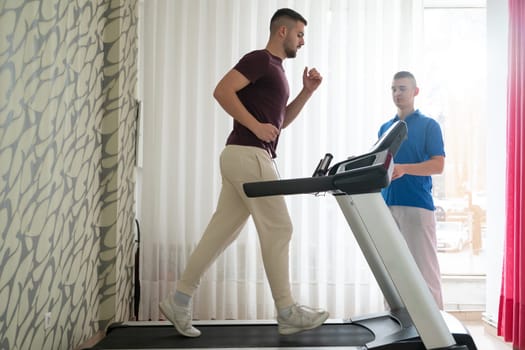 Man undergoing physical therapy with trainer assistance on a treadmill indoors.