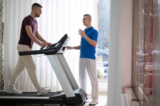 Man undergoing physical therapy with trainer assistance on a treadmill indoors.