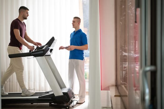 Man undergoing physical therapy with trainer assistance on a treadmill indoors.