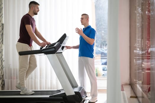 Man undergoing physical therapy with trainer assistance on a treadmill indoors.
