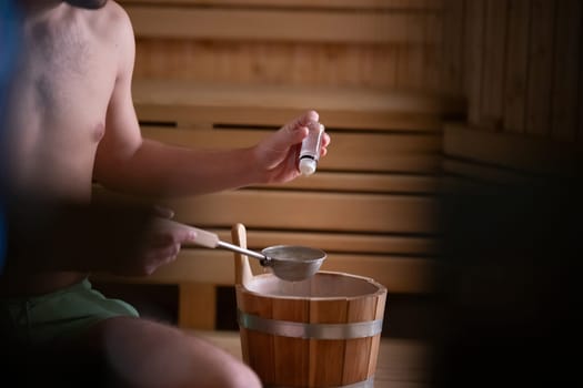 Close up of a mans hand holding essential oils inside a traditional wooden sauna with bucket and ladle.