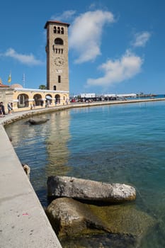 Rodas.Greece - june 04, 2025: Iconic clock tower of Rhodes standing tall by Mandraki Harbour, with serene waters reflecting the vibrant blue sky and white clouds.
