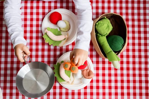Child playing with colorful felt vegetables on red checkered tablecloth.