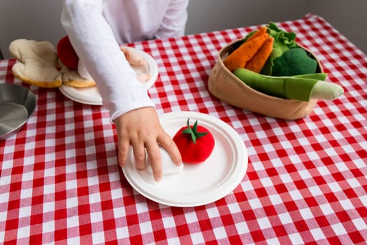 Child playing with toy vegetables on checkered tablecloth.