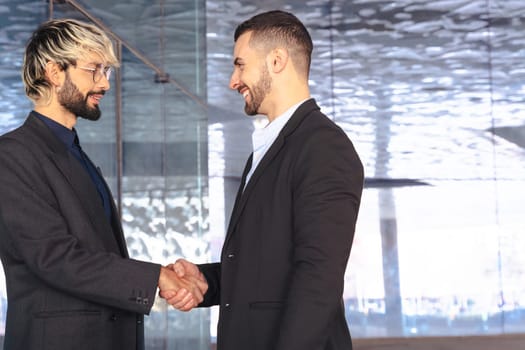 Two professional businessmen in suits shaking hands inside a modern glass office. Business deal, agreement, or partnership in a corporate indoor setting with contemporary design. Copy space