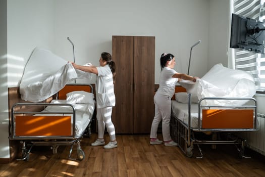 Two nurses in uniforms cleaning and organizing a hospital room by changing the bed linens and ensuring a hygienic environment for patients