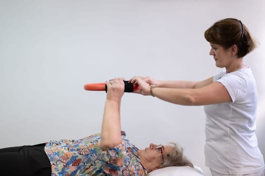 Elderly woman undergoing physiotherapy under the supervision of a female therapist. The patient is lying on a treatment table while performing an arm exercise with an orange pilates ring.