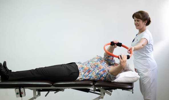 Elderly woman undergoing physiotherapy under the supervision of a female therapist. The patient is lying on a treatment table while performing an arm exercise with an orange pilates ring.