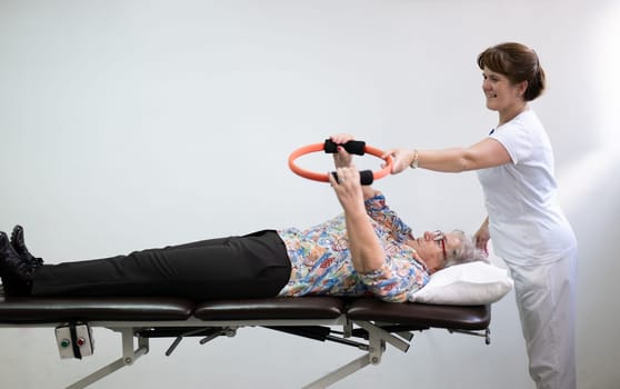 Elderly woman undergoing physiotherapy under the supervision of a female therapist. The patient is lying on a treatment table while performing an arm exercise with an orange pilates ring.