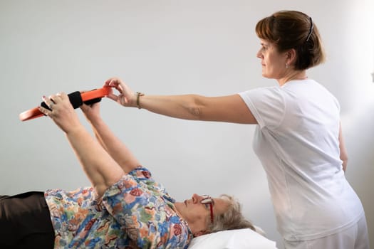 Elderly woman undergoing physiotherapy under the supervision of a female therapist. The patient is lying on a treatment table while performing an arm exercise with an orange pilates ring.
