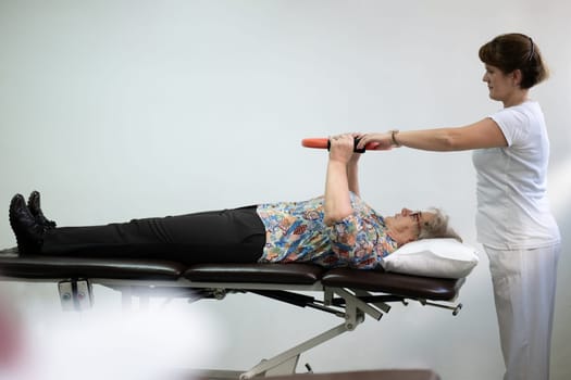 Elderly woman undergoing physiotherapy under the supervision of a female therapist. The patient is lying on a treatment table while performing an arm exercise with an orange pilates ring.