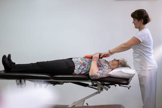 Elderly woman undergoing physiotherapy under the supervision of a female therapist. The patient is lying on a treatment table while performing an arm exercise with an orange pilates ring.