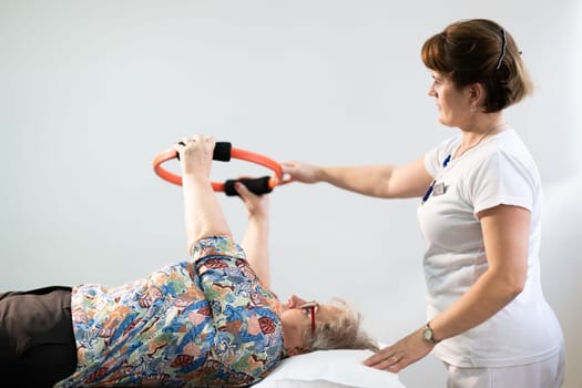 Elderly woman undergoing physiotherapy under the supervision of a female therapist. The patient is lying on a treatment table while performing an arm exercise with an orange pilates ring.