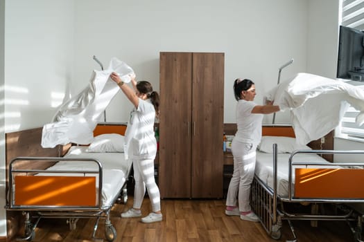 Two nurses in uniforms cleaning and organizing a hospital room by changing the bed linens and ensuring a hygienic environment for patients