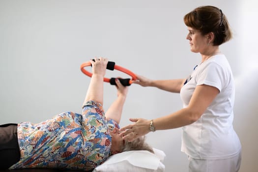 Elderly woman undergoing physiotherapy under the supervision of a female therapist. The patient is lying on a treatment table while performing an arm exercise with an orange pilates ring.