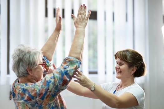 Senior woman doing physical exercises with the help of a smiling therapist. Concept of elderly rehabilitation and movement recovery.