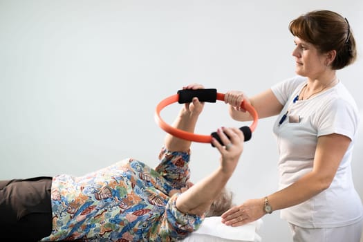 Elderly woman undergoing physiotherapy under the supervision of a female therapist. The patient is lying on a treatment table while performing an arm exercise with an orange pilates ring.