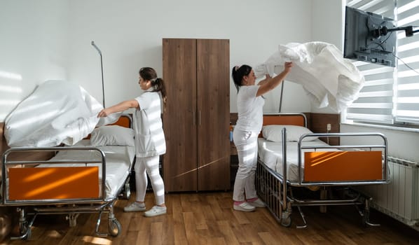 Two nurses in uniforms cleaning and organizing a hospital room by changing the bed linens and ensuring a hygienic environment for patients