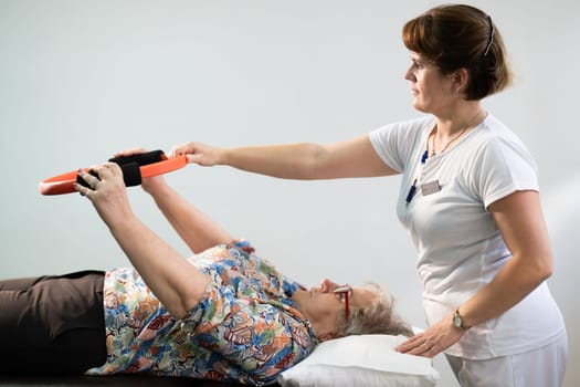 Elderly woman undergoing physiotherapy under the supervision of a female therapist. The patient is lying on a treatment table while performing an arm exercise with an orange pilates ring.