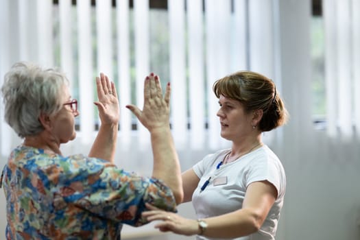 Senior woman doing physical exercises with the help of a smiling therapist. Concept of elderly rehabilitation and movement recovery.