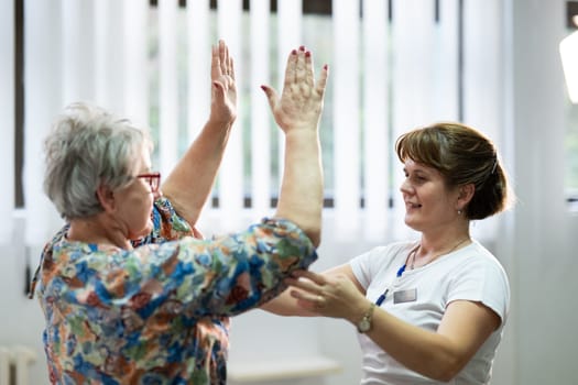 Senior woman doing physical exercises with the help of a smiling therapist. Concept of elderly rehabilitation and movement recovery.