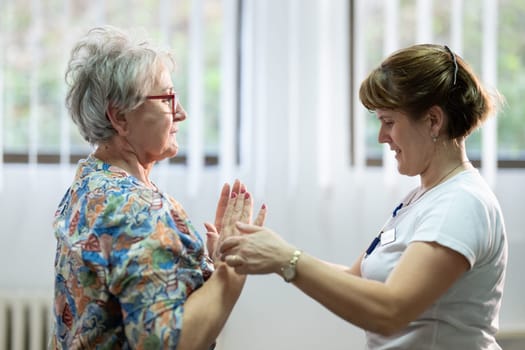 Senior woman doing physical exercises with the help of a smiling therapist. Concept of elderly rehabilitation and movement recovery.