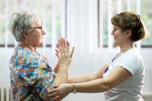 Senior woman doing physical exercises with the help of a smiling therapist. Concept of elderly rehabilitation and movement recovery.