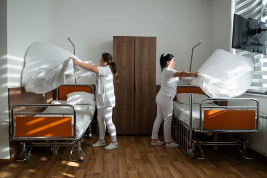 Two nurses in uniforms cleaning and organizing a hospital room by changing the bed linens and ensuring a hygienic environment for patients