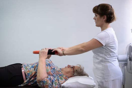 Elderly woman undergoing physiotherapy under the supervision of a female therapist. The patient is lying on a treatment table while performing an arm exercise with an orange pilates ring.