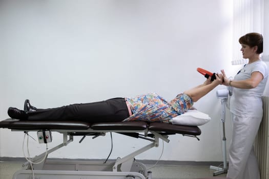 Elderly woman undergoing physiotherapy under the supervision of a female therapist. The patient is lying on a treatment table while performing an arm exercise with an orange pilates ring.