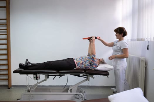 Elderly woman undergoing physiotherapy under the supervision of a female therapist. The patient is lying on a treatment table while performing an arm exercise with an orange pilates ring.