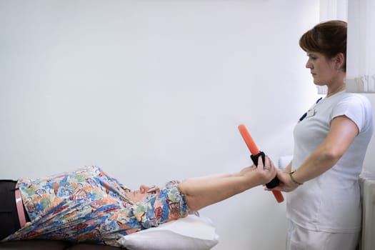 Elderly woman undergoing physiotherapy under the supervision of a female therapist. The patient is lying on a treatment table while performing an arm exercise with an orange pilates ring.