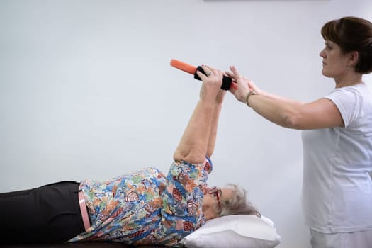 Elderly woman undergoing physiotherapy under the supervision of a female therapist. The patient is lying on a treatment table while performing an arm exercise with an orange pilates ring.
