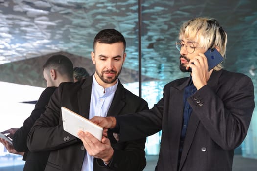 Two young businessmen in suits collaborating in a modern office. One uses a tablet while the other speaks on the phone, surrounded by reflective glass and a stylish professional atmosphere.