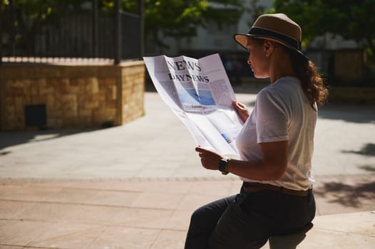 A woman sits outdoors in the sunlight, reading a newspaper. She wears a hat and casual attire, creating a peaceful and relaxed scene amidst warm ambient lighting.