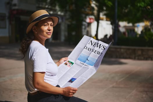 A woman wearing a hat reads a newspaper while sitting relaxed outdoors in a park on a sunny day, enjoying the fresh air and tranquility.