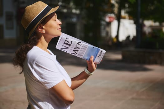 A woman wearing a straw hat and white shirt enjoys a sunny day outdoors, holding a folded newspaper in a serene urban street setting.