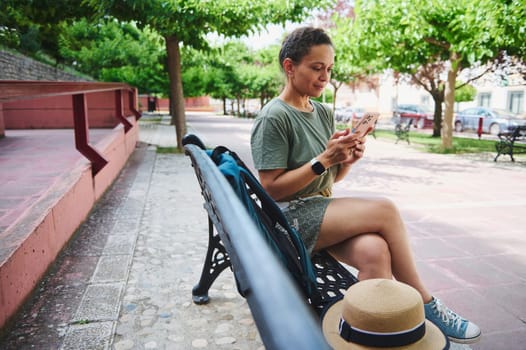 Individual seated on a park bench in a tranquil outdoor environment while interacting with their smartphone. Capturing a moment of relaxation and connectivity amidst natural surroundings.