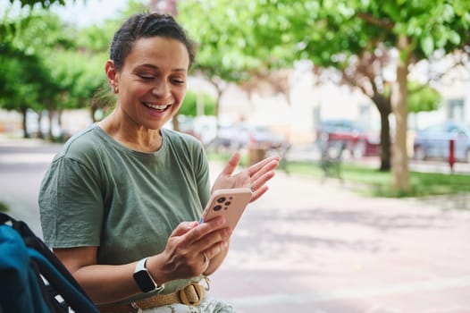 Smiling woman holding a smartphone, enjoying a conversation in a sunny park. Leans forward animatedly, expressing joy and engagement during the virtual interaction.