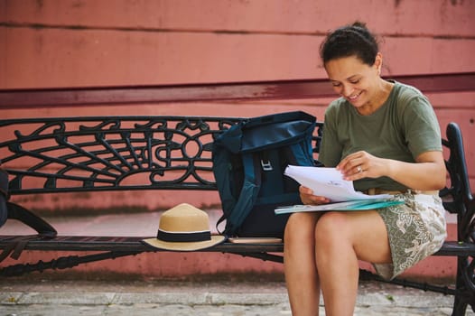 Smiling woman sitting on a decorative bench, reading documents with an open backpack and a straw hat nearby, enjoying the sunny outdoor setting, relaxed and immersed in her activity.