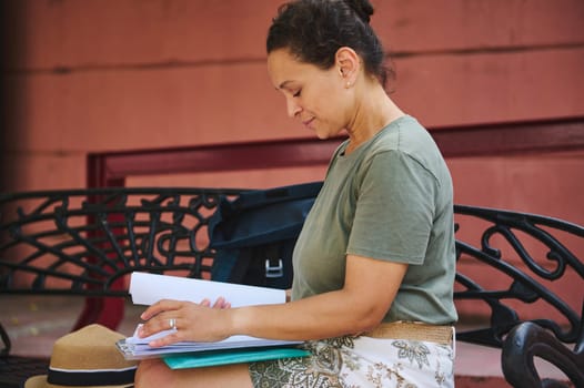 A woman sits on a decorative park bench reading through papers. The scene reflects focus and tranquility, emphasizing outdoor leisure with nearby belongings.