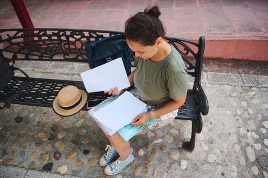A woman sits on a decorative bench outdoors, examining documents. The scene conveys focus and tranquility, with accessories like a hat and backpack enhancing the casual ambiance.