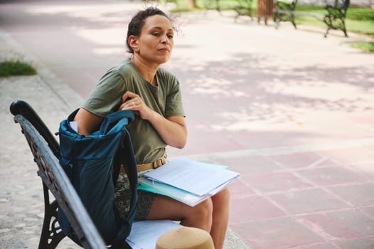 A woman sits on a park bench, organizing papers and items from her backpack, enjoying the relaxed outdoor setting on a sunny day.