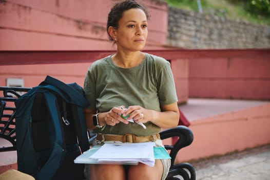 A woman sits outdoors on a bench, holding papers and a pen, immersed in thought. She appears to be analyzing or planning with focus during outdoor activities.